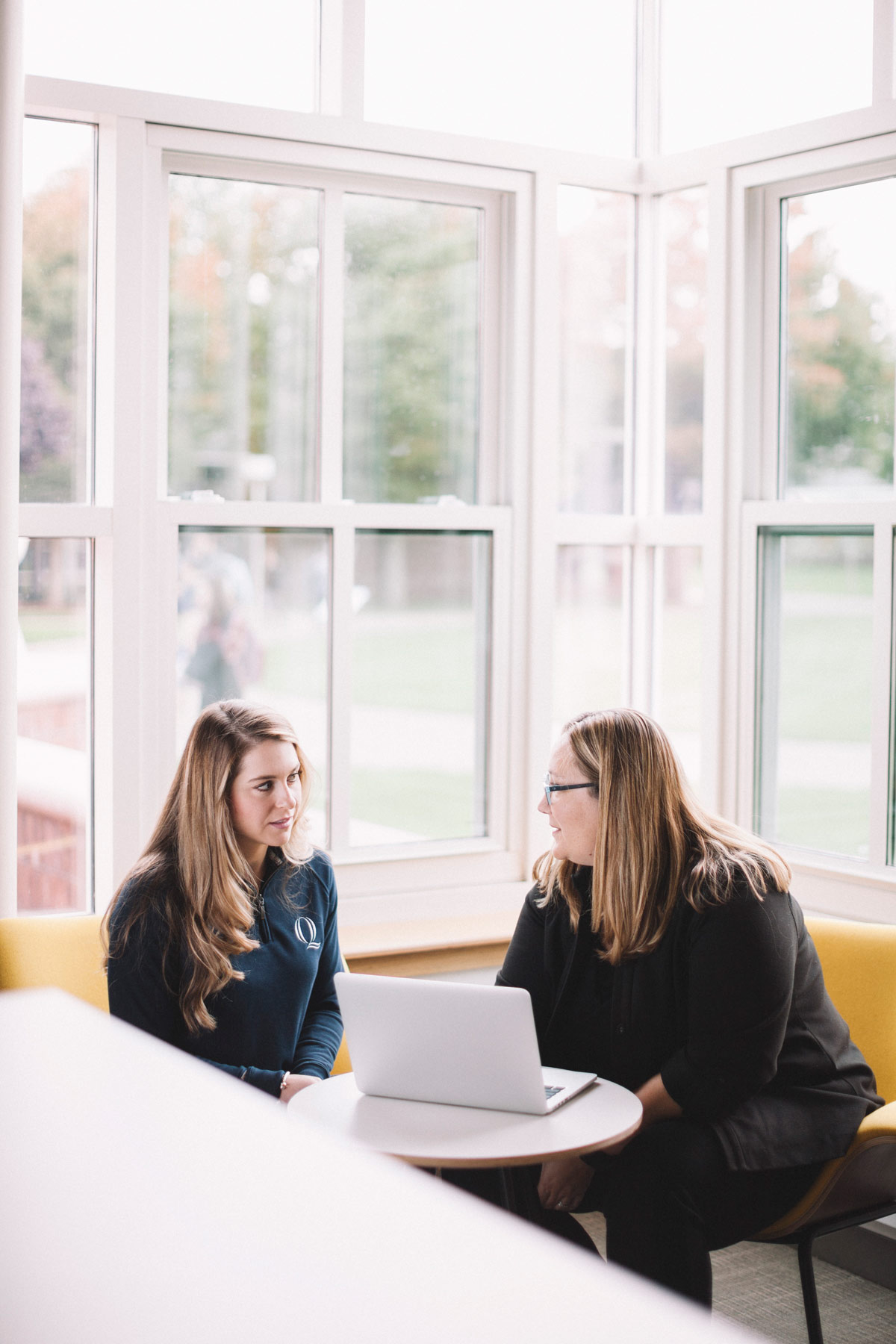 Jill Koehler sits and talks with a student at a table with a laptop surrounded by windows looking onto the quad.